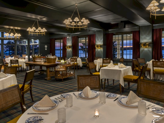 The image shows an elegant restaurant dining area with neatly set tables, chandeliers, and large windows, offering a view outside during twilight.