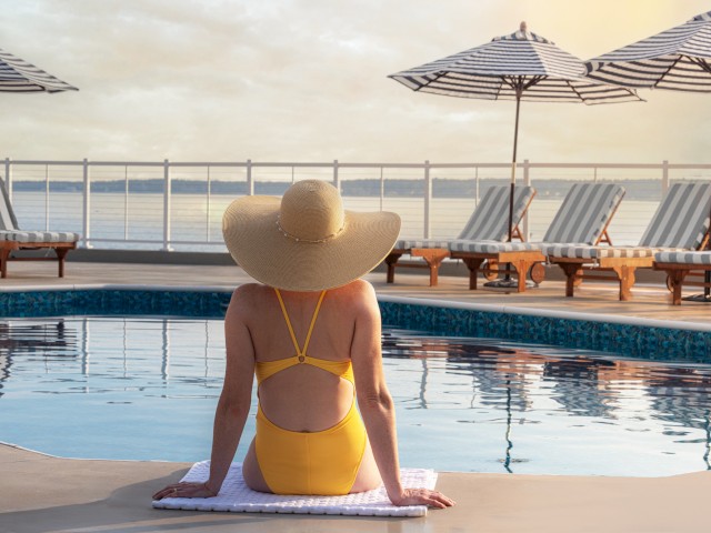 A person in a yellow swimsuit and sunhat sits poolside on a mat, gazing at the water, with loungers and umbrellas in the background.