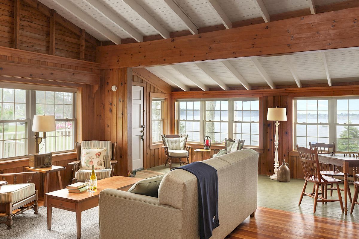 A cozy living room with wooden walls, large windows, and a mix of armchairs, a sofa, and a dining table. Natural light fills the space.