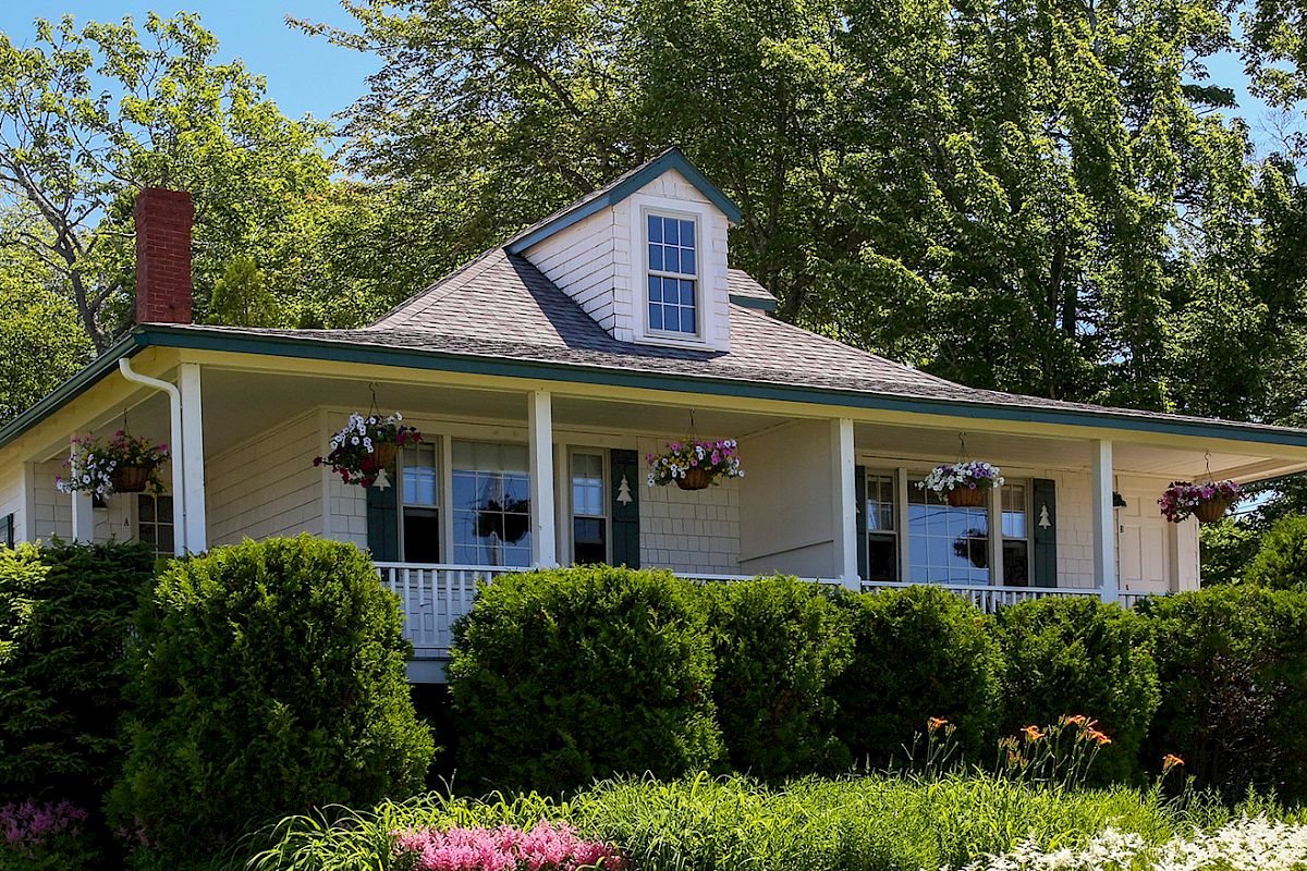 A charming house with a porch, hanging flower baskets, surrounded by lush shrubs and colorful flowers, nestled among trees under a clear blue sky.