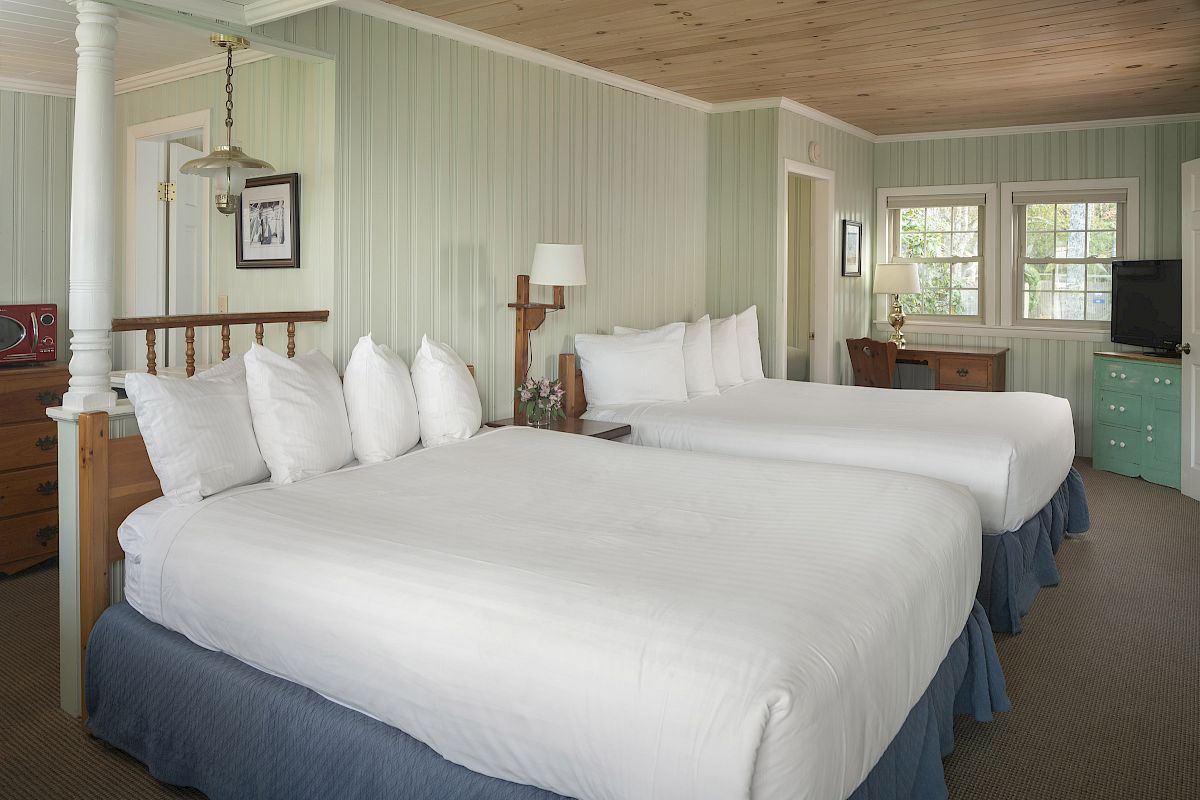 A cozy hotel room with light green walls, two double beds, a flat-screen TV, a desk, and white linens, decorated in a country style.