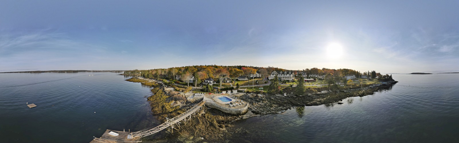 Aerial view of a scenic coastal area with a pier, rocky shore, houses, and lush trees under a bright sky, and the sun low in the sky, ending the sentence.