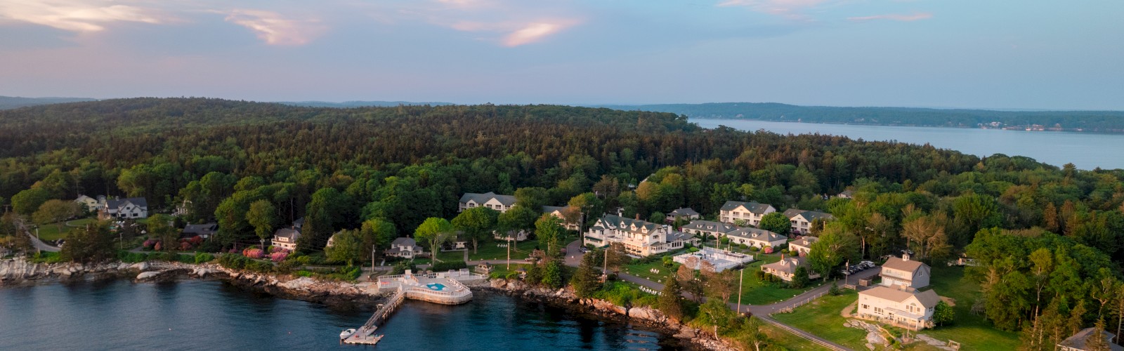 Aerial view of a coastal area with houses, forest, and a dock jutting into calm water under a partly cloudy sky.