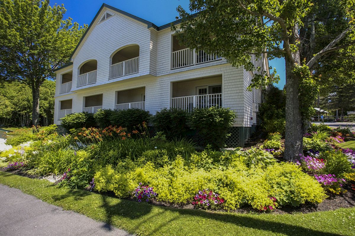 A white two-story house with balconies, surrounded by lush greenery, colorful flowers, and a tree on a sunny day.