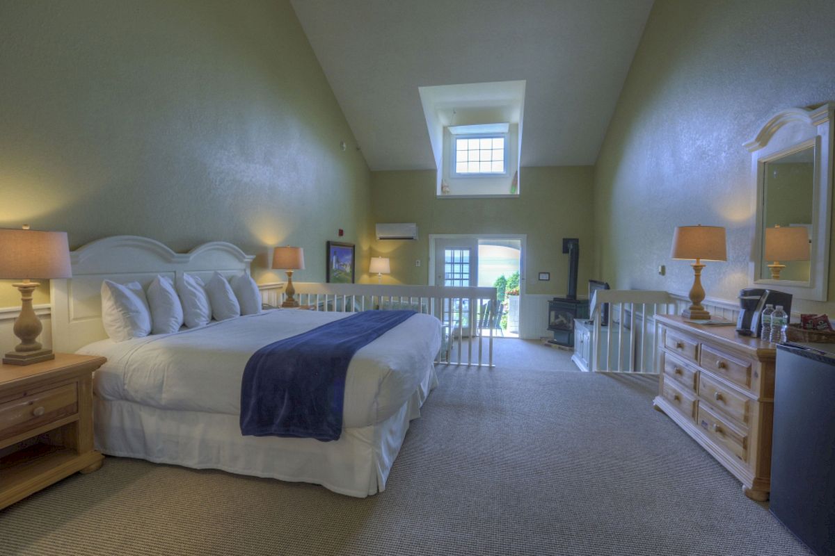 A bedroom with a large bed, nightstands, lamps, dresser with mirror, skylight, and balcony overlooking greenery, all in a spacious setting.