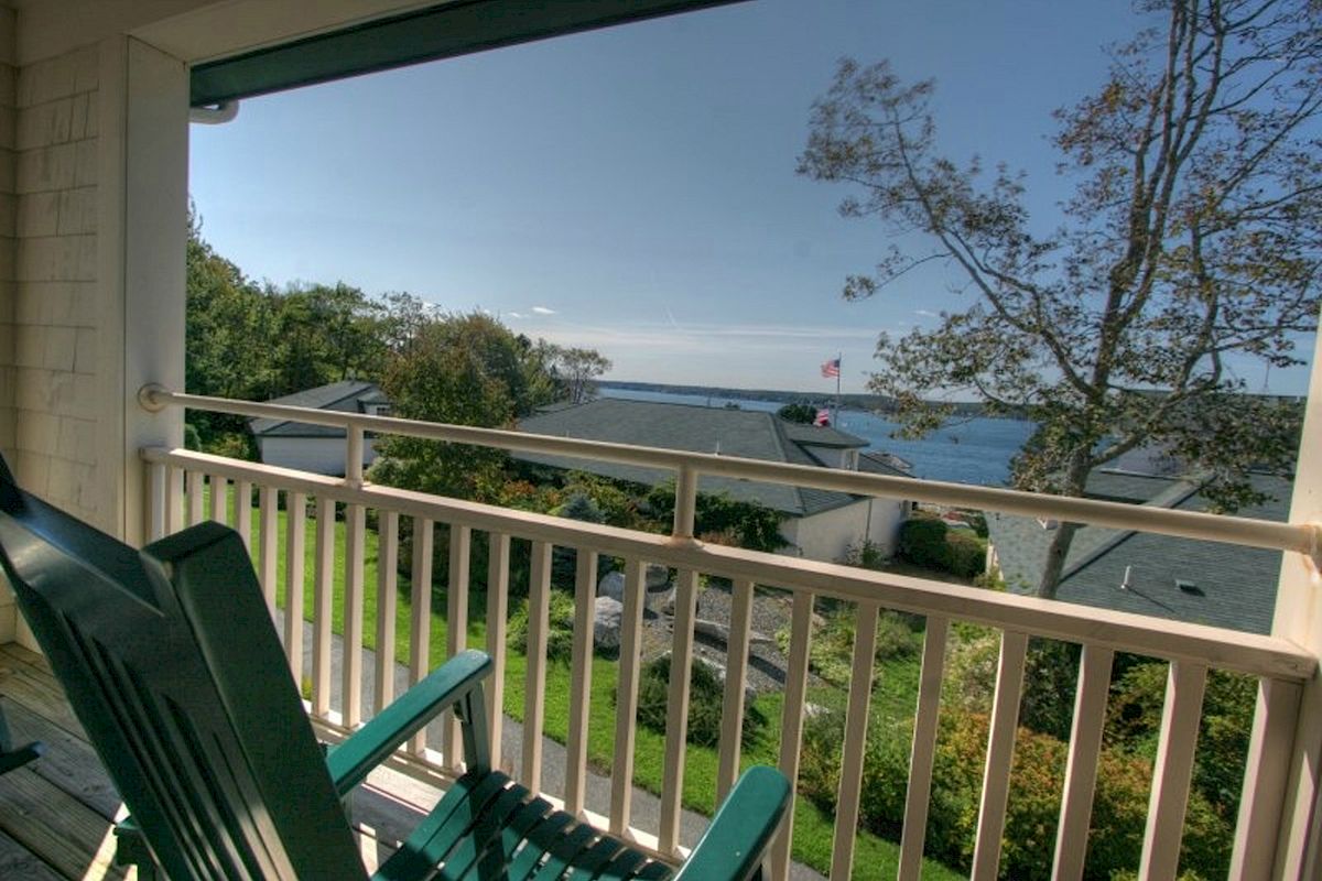 Two green chairs on a balcony overlook a scenic view of houses, trees, and a body of water under a clear blue sky.