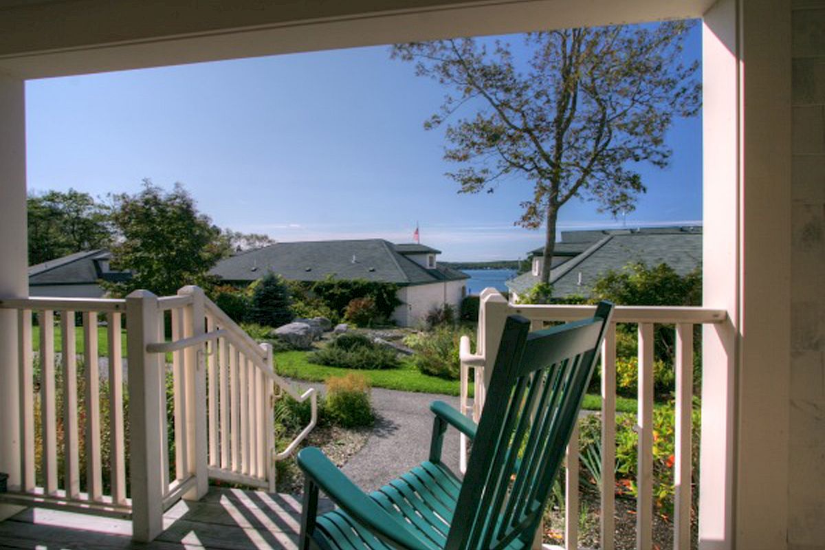 A green rocking chair on a porch overlooks a well-kept garden and buildings with a glimpse of the blue sky and sea in the background.