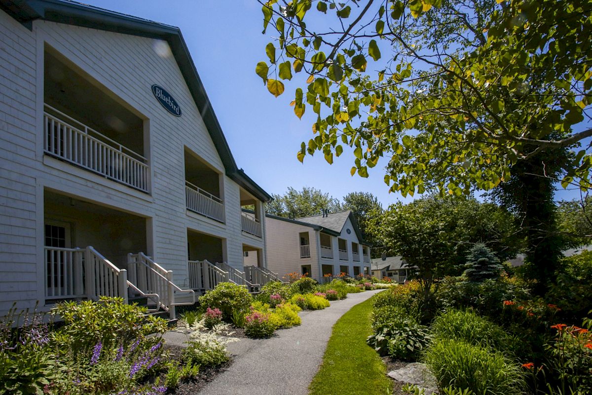 A serene outdoor path with landscaped gardens leads to a residential building with balconies. Trees and flowers enhance the peaceful ambiance.