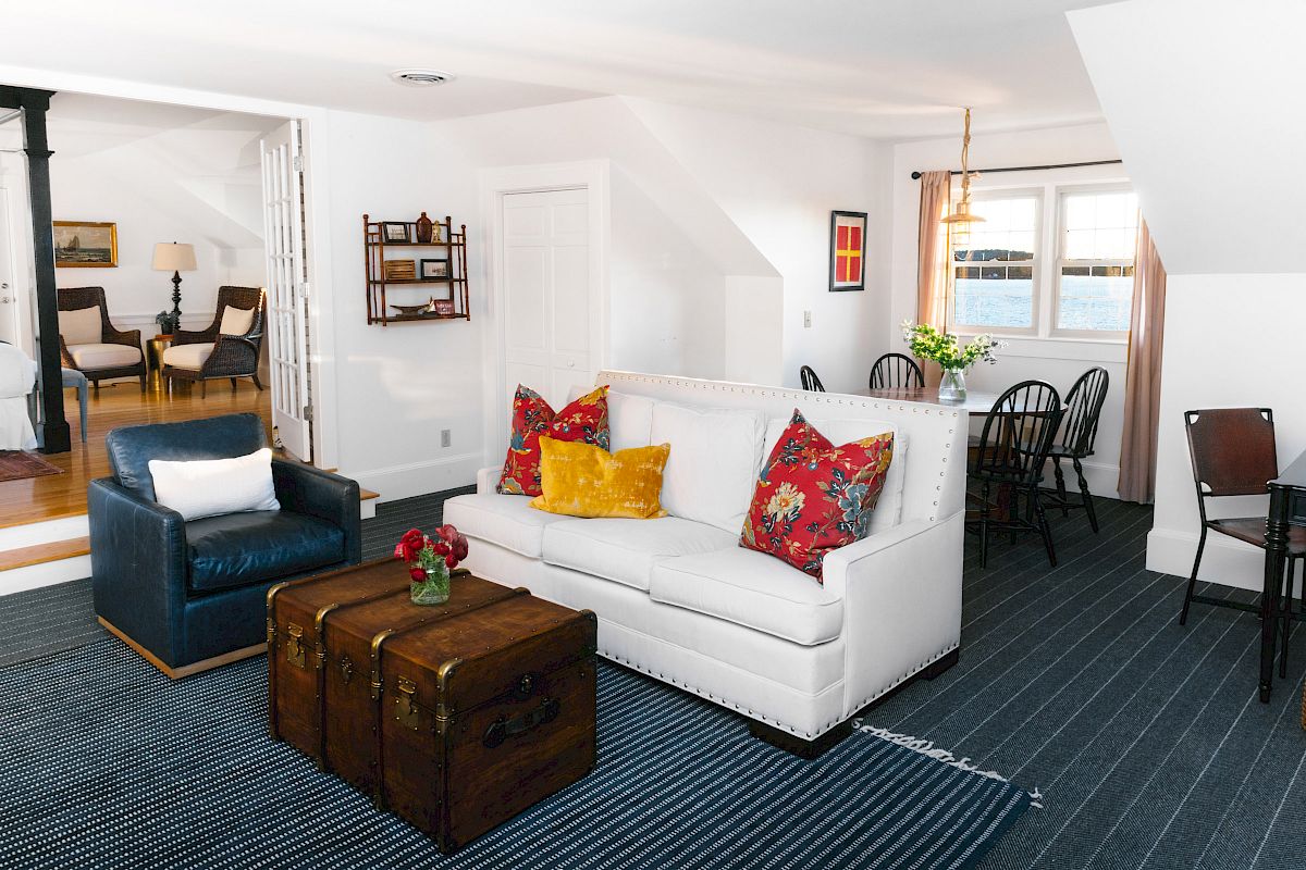 A cozy living room with a white couch, colorful pillows, a trunk coffee table, and a blue armchair near a dining area with dark wood furniture.