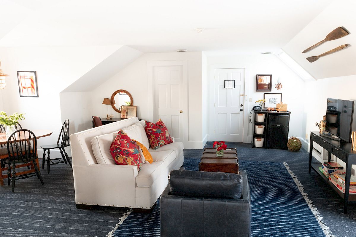 A cozy living space with a white sofa, colorful cushions, a small dining area, and wall art. A TV stand and storage units are also present.