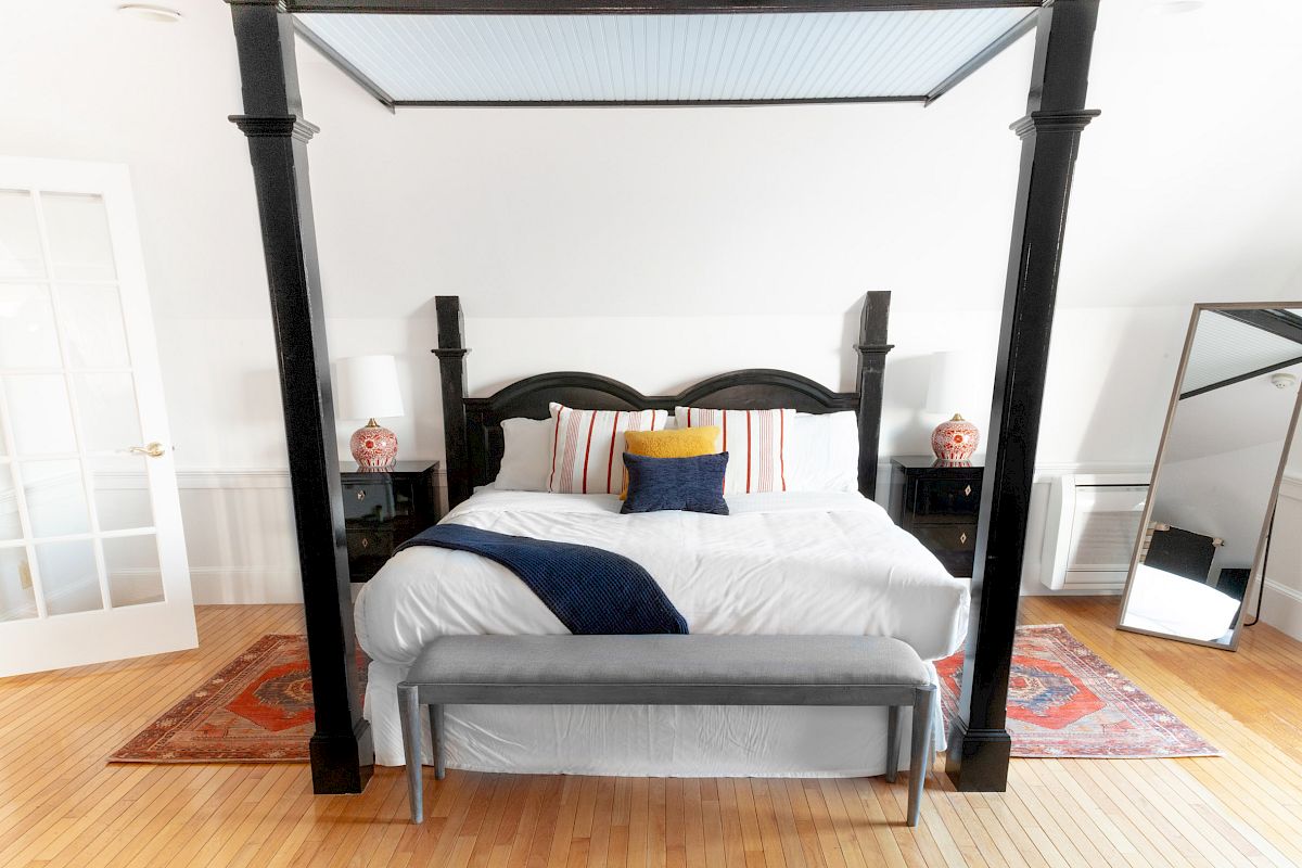 A neatly arranged bedroom with a four-poster bed, white bedding, a blue throw, a bench, two nightstands with lamps, and a large mirror.