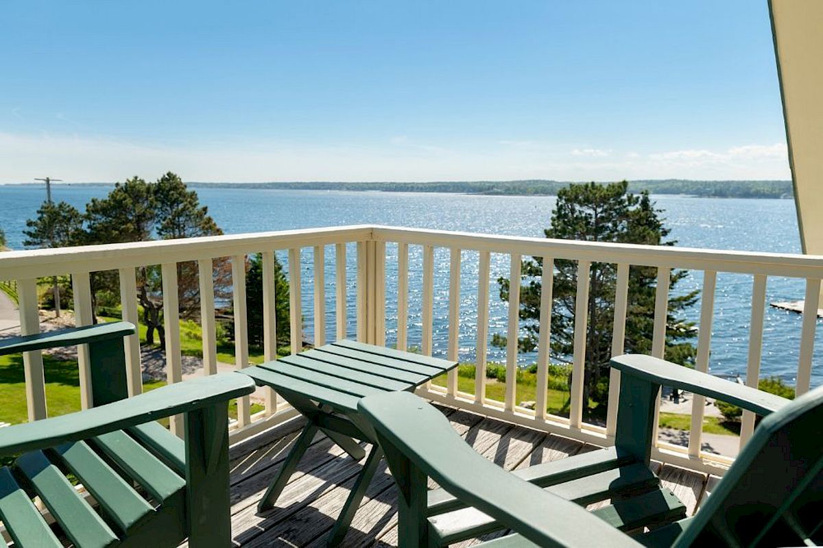 A balcony with green Adirondack chairs overlooks a serene body of water and trees under a clear blue sky.