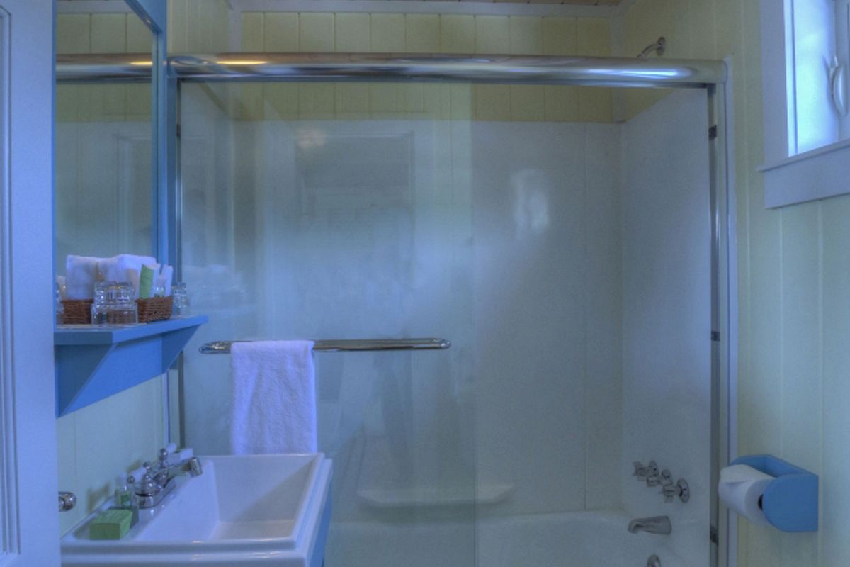 A bathroom with a shower-bathtub combo, blue vanity with sink, a wall mirror, wooden ceiling, towel rack, toilet, and a window above the toilet.
