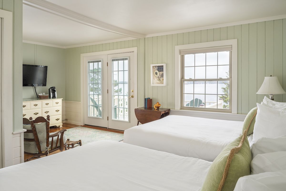 A cozy, well-lit room with two beds, a TV, a desk, and a dresser. The room has windows with a view and double doors opening to the outside.