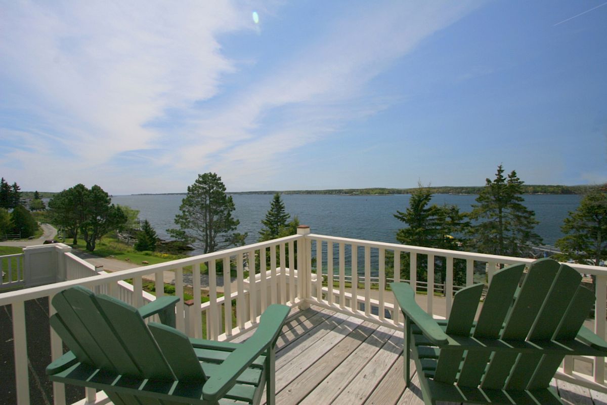 The image shows a balcony with two green Adirondack chairs overlooking a scenic lake with trees and a clear blue sky in the background.