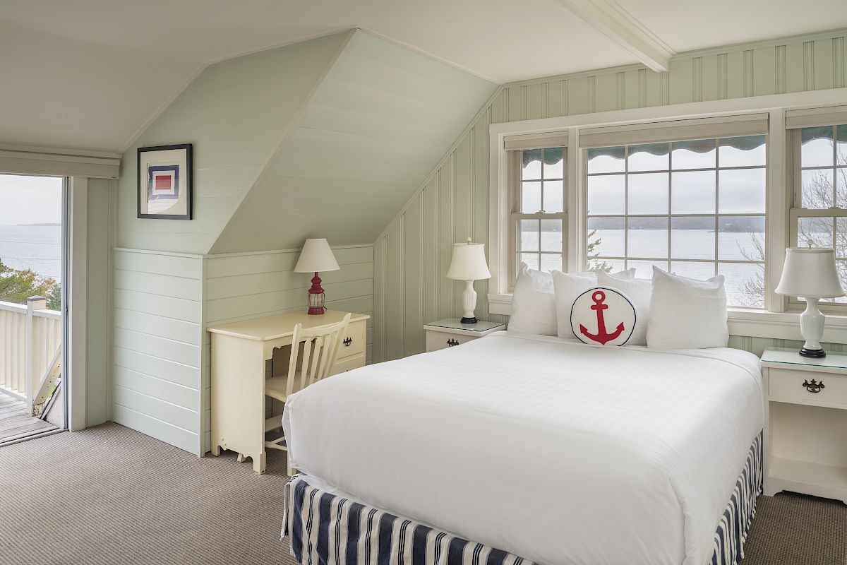 A cozy bedroom with a white bed, nautical-themed pillow, desk, two lamps, a window with an ocean view, and light green walls with slanted ceiling.