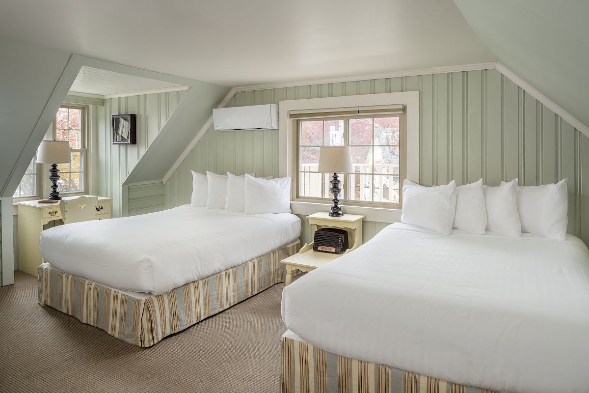 A cozy room with two double beds, nightstands with lamps, a window, a dresser with a TV, pillows, an air conditioning unit, and light green walls.