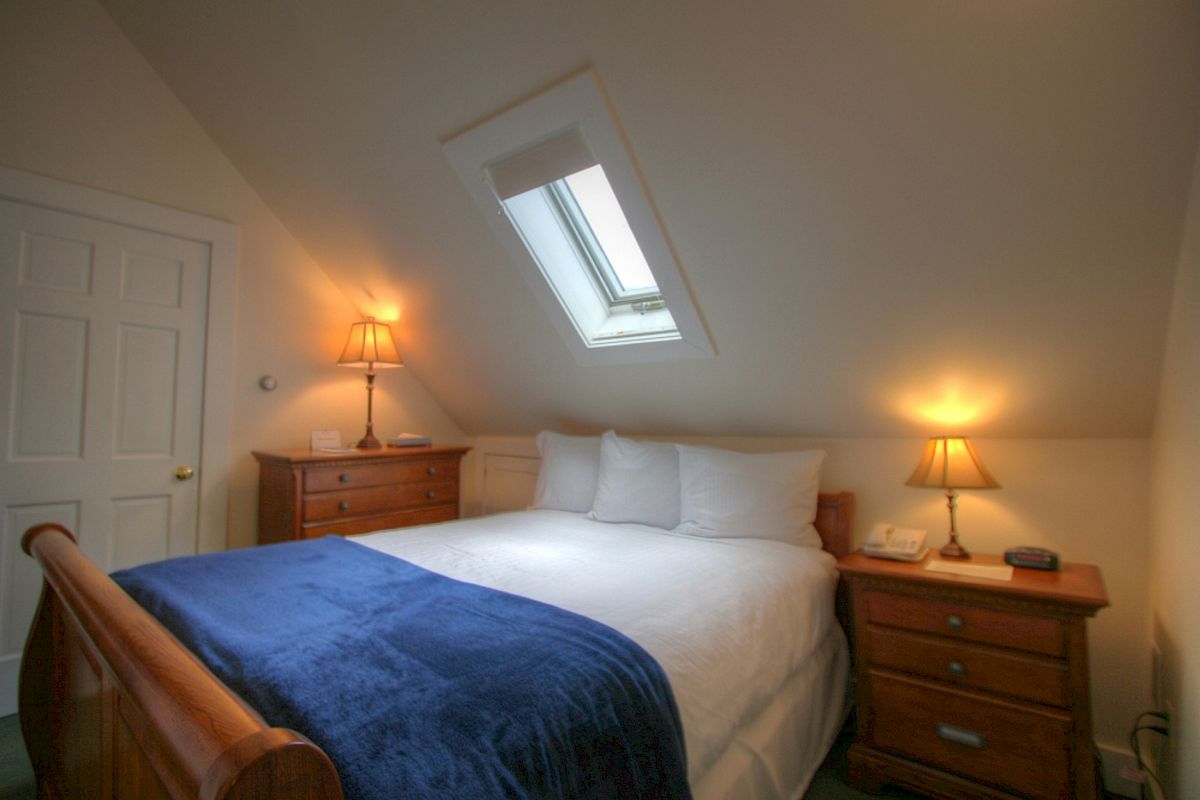 A cozy bedroom with a slanted ceiling, a skylight, a double bed with white linens and a blue throw, and lamps on wooden nightstands, ending the sentence.