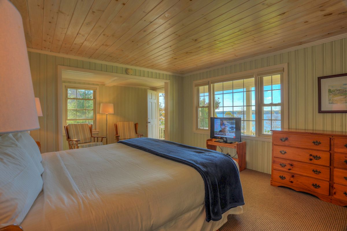 A cozy bedroom with a wooden ceiling, a bed with a blue throw, a TV on a dresser, and two chairs by the window.