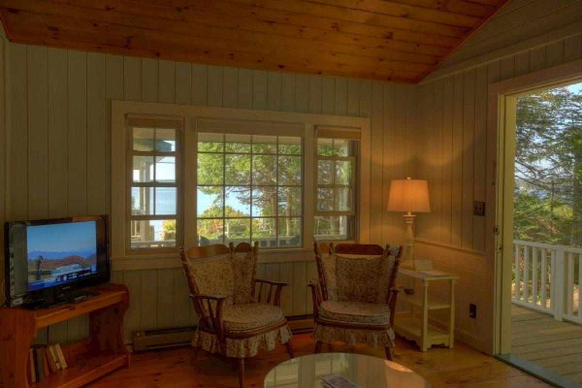 A cozy room with wooden interiors, two armchairs, a TV, and a lamp. A window and door offer views of trees and outdoor scenery, ending the sentence.