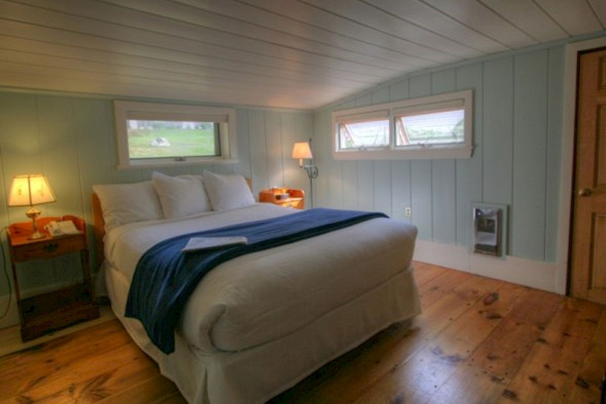 A cozy bedroom with wooden floors, a neatly made bed, blue blanket, bedside tables, lamps, and small windows letting in natural light.
