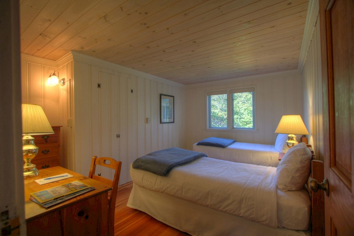The image shows a cozy bedroom with two single beds, a desk, a chair, lamps on nightstands, a wooden ceiling, and windows letting in natural light.