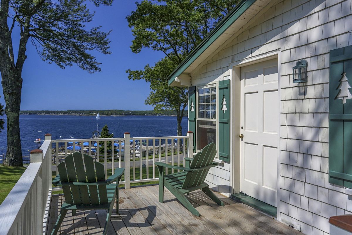 This image shows a porch with two Adirondack chairs facing a scenic lake or bay, adjacent to a house with green shutters and surrounded by trees.