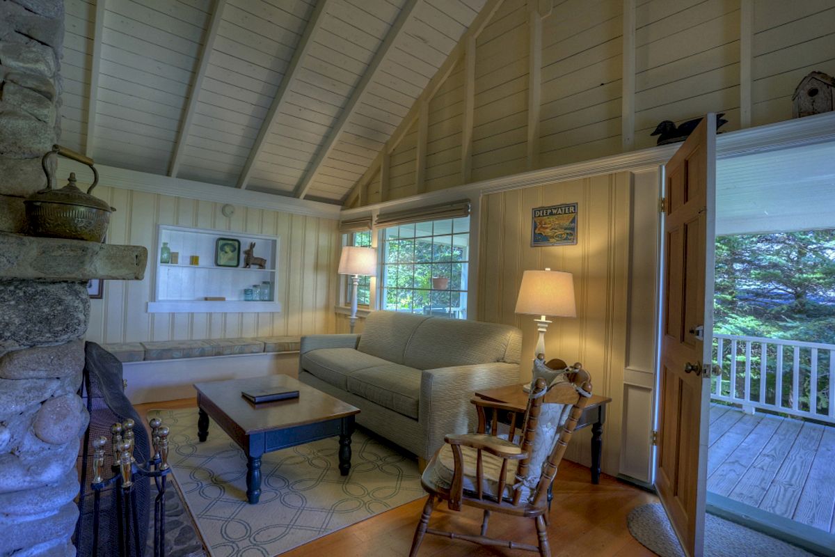 This image shows a cozy living room with a high, vaulted ceiling, a sofa, a coffee table, lamps, and a rocking chair, with an open door leading outside.