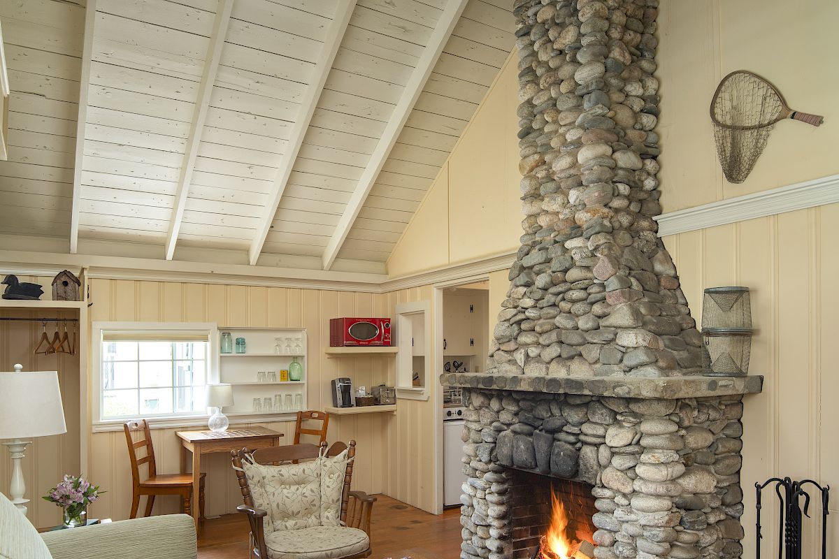 This image shows a cozy living room with a high wooden ceiling, a stone fireplace, a sofa, a coffee table, and a small dining area.