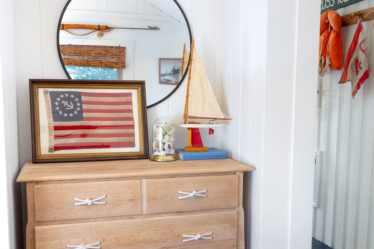 Wooden dresser with four drawers, a round mirror, framed American flag, and decorative sailboat. Wall-mounted lamp above. Room visible on the right.