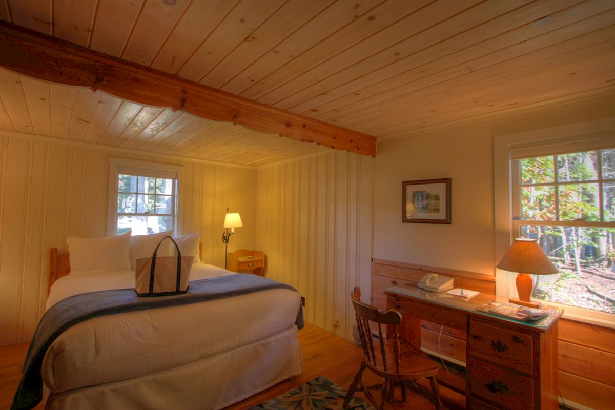 A cozy bedroom with wooden walls and ceiling, a bed with white linens, a desk with a lamp, and two windows letting in natural light.