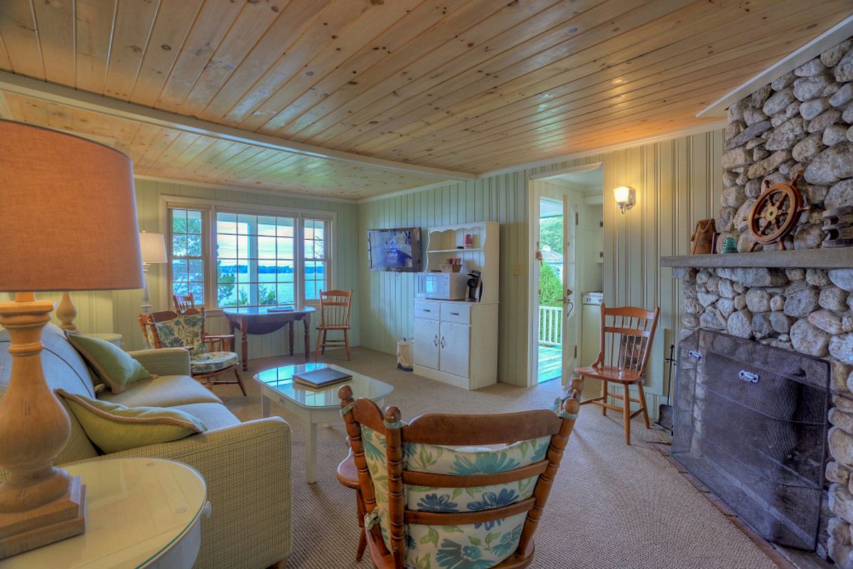 A cozy living room with wooden ceiling, stone fireplace, cushioned seating, a TV, and large windows letting in natural light, ending the sentence.