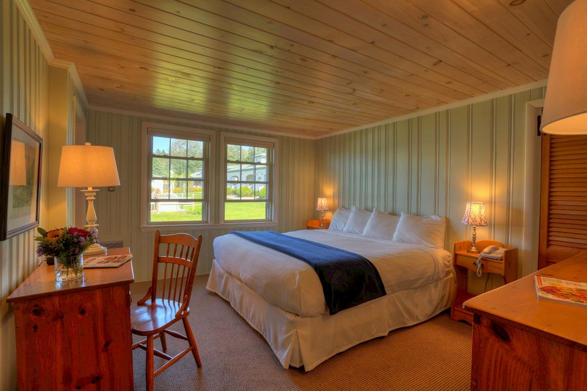 A cozy bedroom with a large bed, wooden furniture, and a desk. It has windows letting in natural light and is brightly lit with lamps.