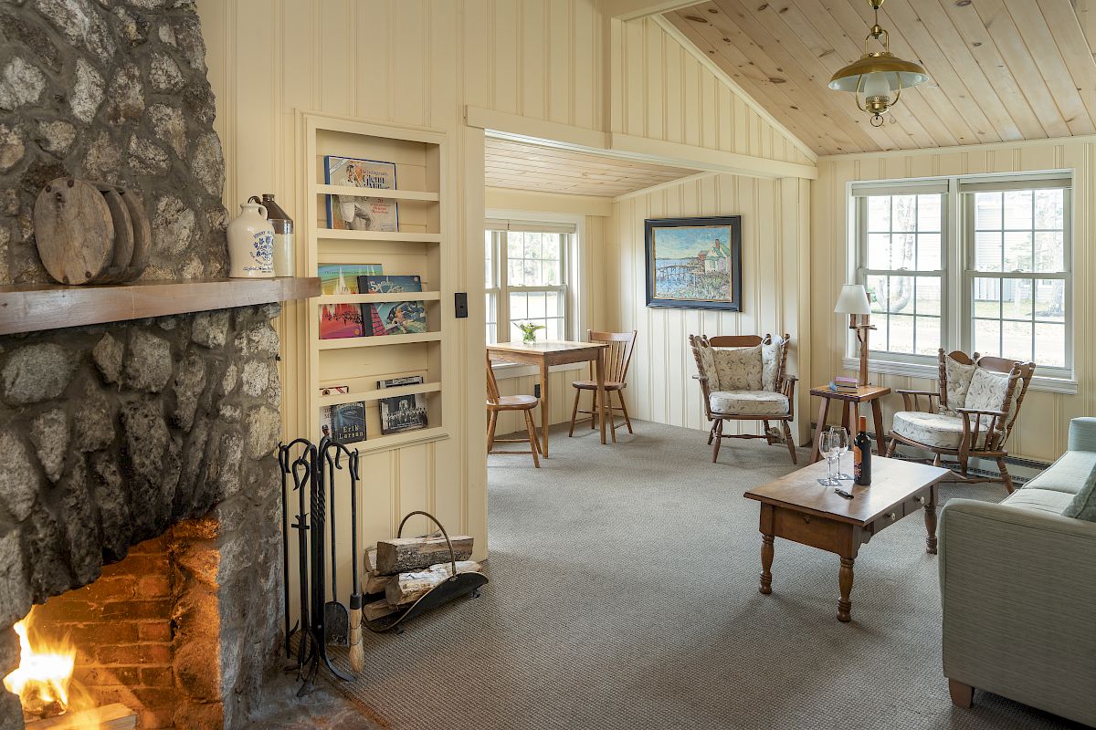 A cozy living room with a stone fireplace, bookshelves, seating area, a wooden coffee table, windows letting in natural light, and a small dining area.