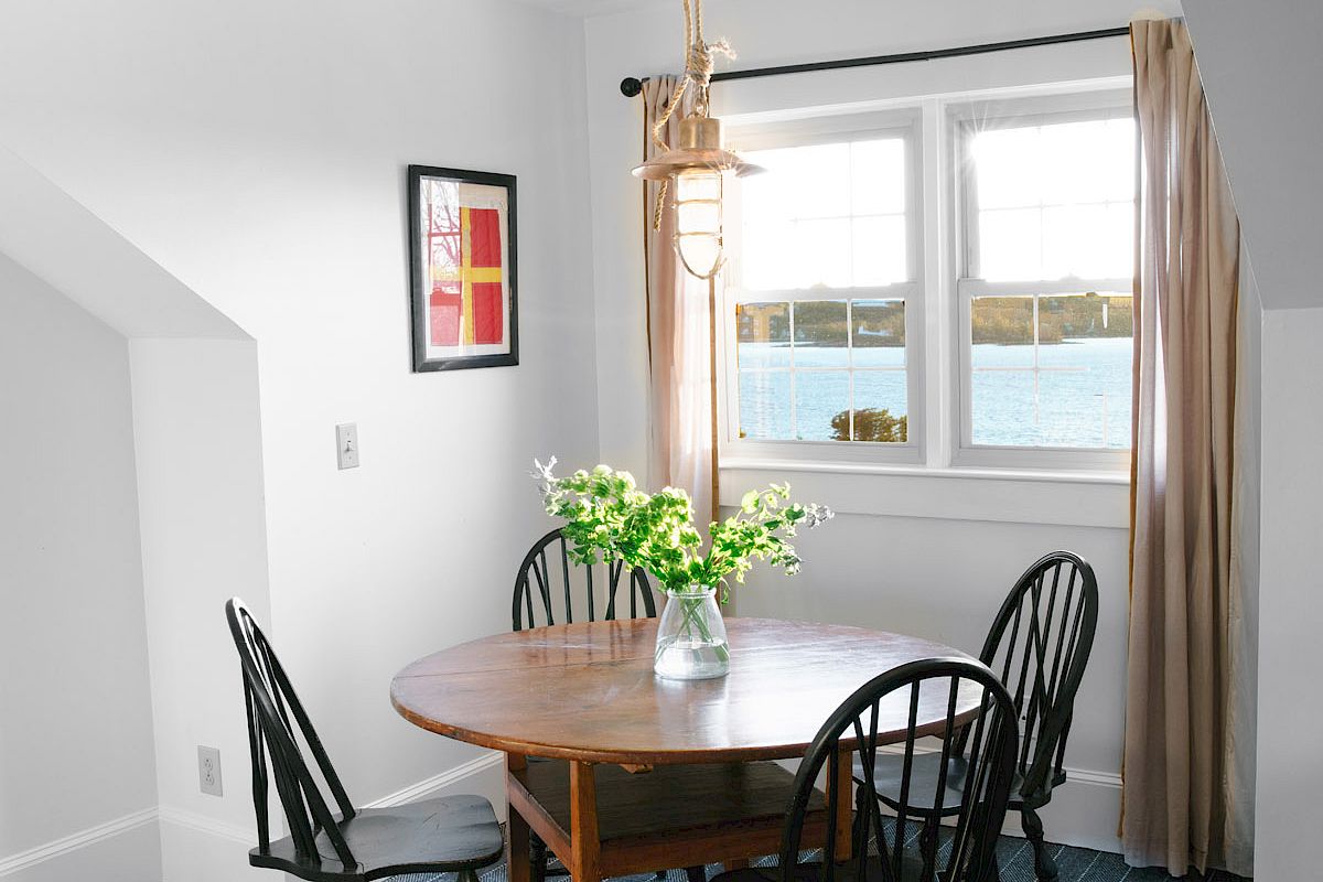 A cozy dining area with a round table, five black chairs, a vase with flowers, a window with a view, a pendant light, and wall art.