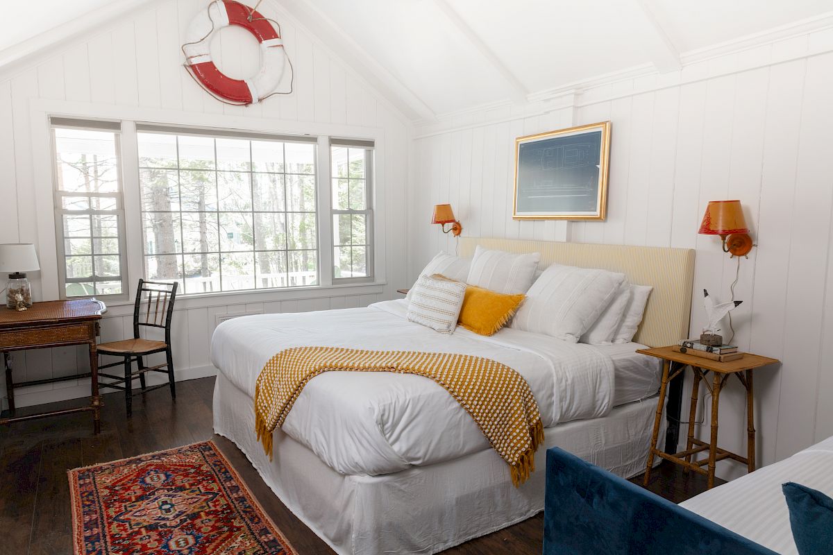 A cozy bedroom with a large bed, a yellow throw blanket, side tables, a desk with a chair, a lifebuoy on the wall, and a window.
