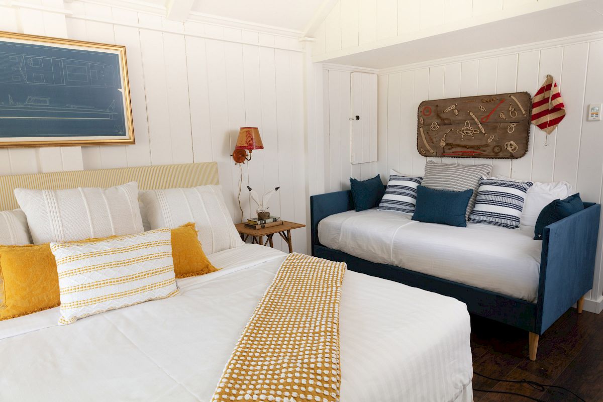 A cozy bedroom with a bed, a daybed, pillows in white and blue, yellow blankets, and a nautical-themed decor mounted on the wall.