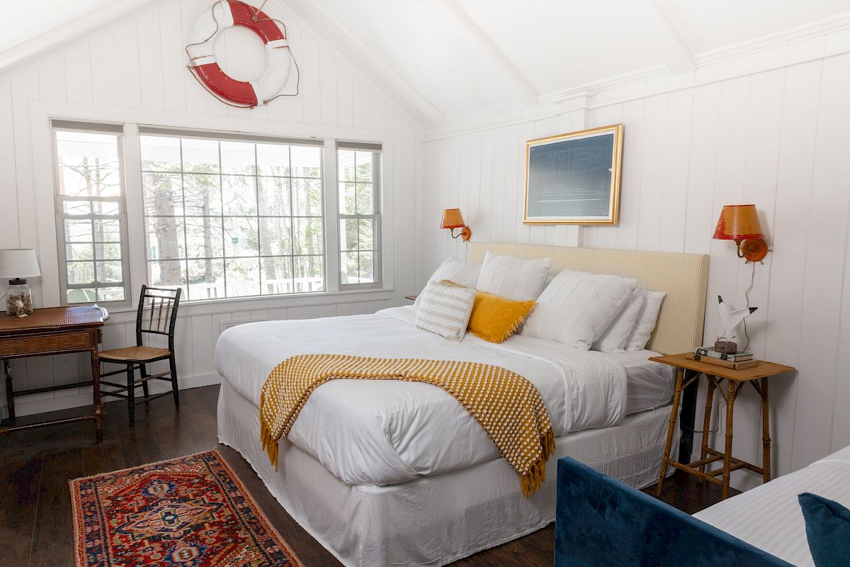 A cozy bedroom with a large bed, yellow pillow, and knitted throw. A window, life preserver decor, desk, chair, and side tables complete the look.