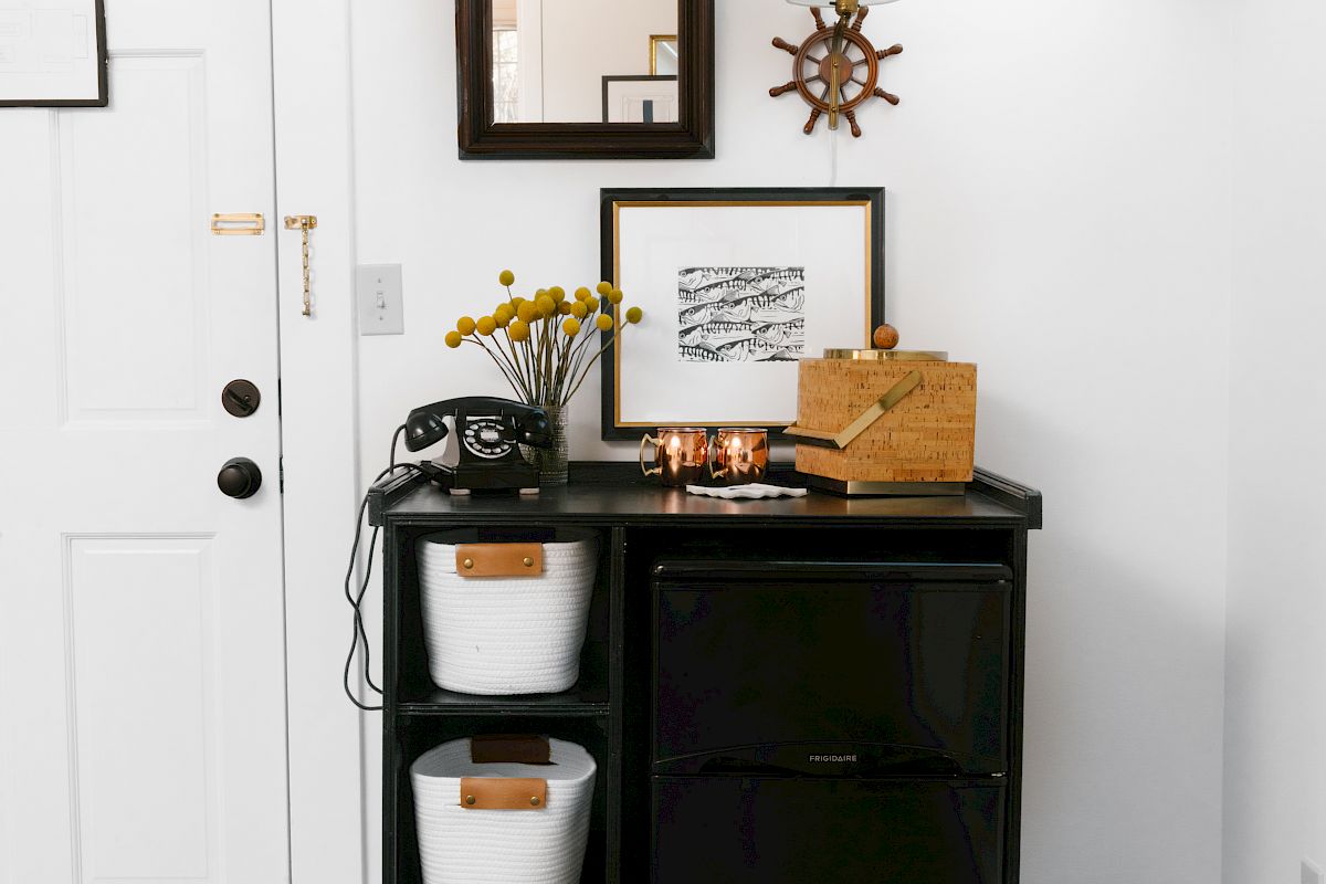A small black storage unit with white bins, a phone, and decor items, including framed pictures, a mirror, and a lamp on a white wall.