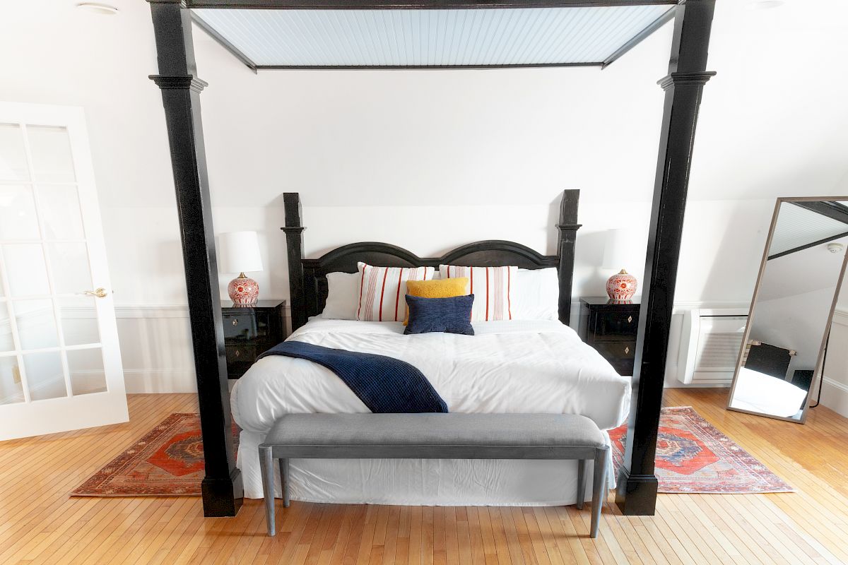 A bedroom with a four-poster bed, a cushioned bench at the foot, two bedside tables with lamps, a rug, a mirror, and a door with glass panes.