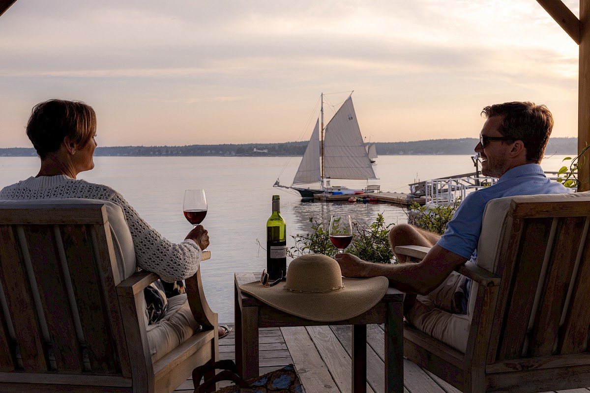 Two people are sitting on a deck with wine glasses, overlooking a peaceful lake with a sailboat in the distance, at sunset.