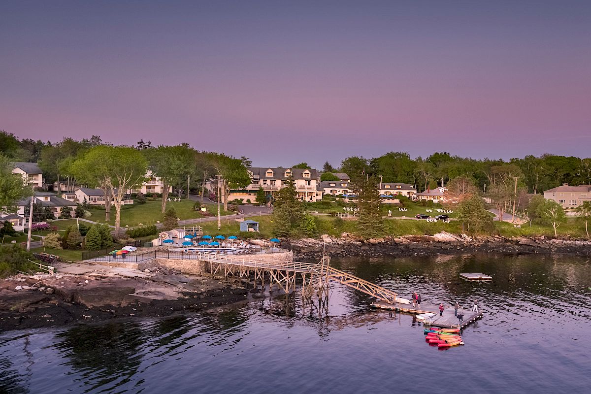 The image shows a coastal village at dusk with houses, a dock, and kayaks on calm water. The sky has a gradient of pastel colors ending the sentence.