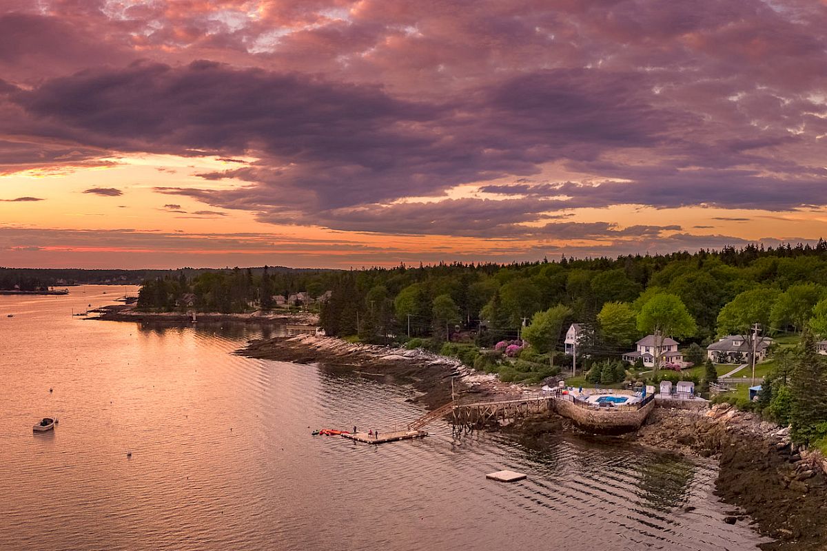 A picturesque coastal village at sunset with sailboats on the water, houses nestled among trees, and a vibrant, colorful sky above.