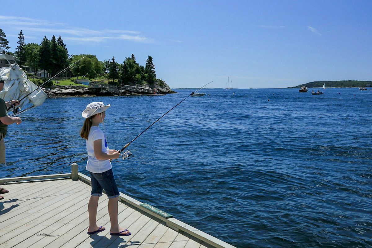 A child and an adult are fishing off a wooden dock by the sea with boats and an island in the background on a sunny day.