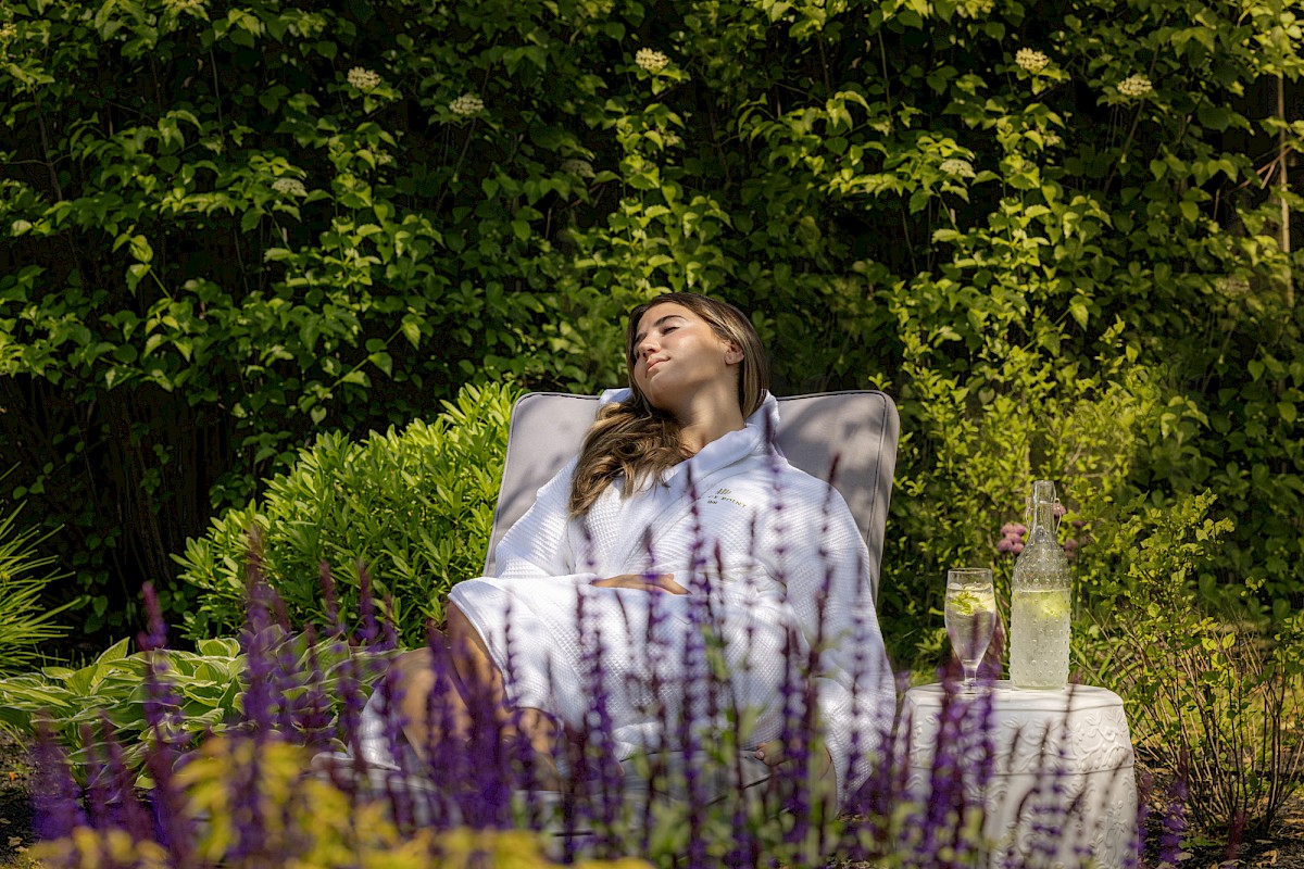 A woman is relaxing on a lounge chair in a garden, surrounded by greenery and flowers, with a drink on a small table beside her.