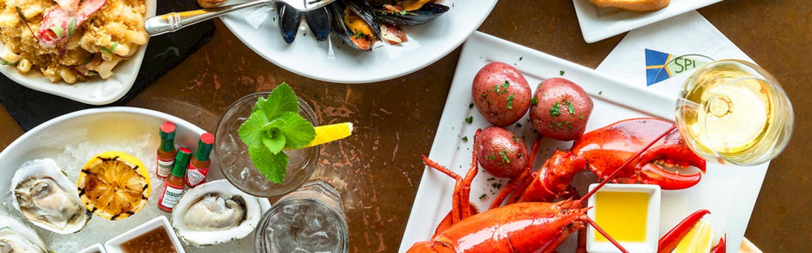 The image shows a variety of seafood dishes including lobster, oysters, mussels, and grilled bread, along with drinks and condiments on a table.
