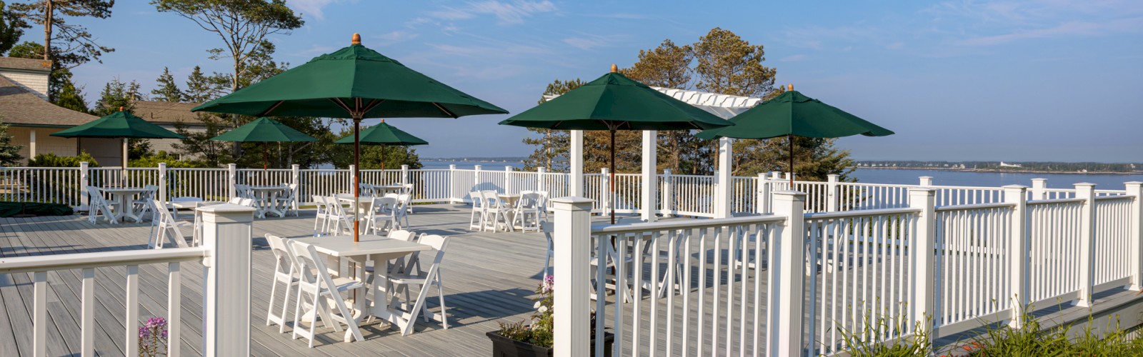 Outdoor deck with white railings, tables, and green umbrellas overlooking a scenic water view, surrounded by trees and flowers.