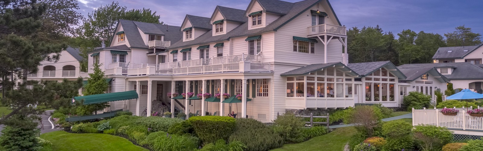 This image shows a large, multi-story house with a manicured lawn, white Adirondack chairs, and a backdrop of trees under a purple sky.