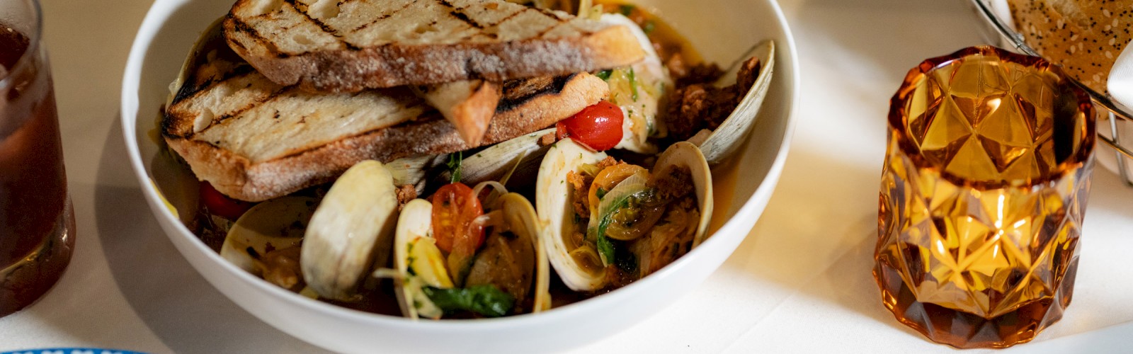 A bowl of seafood pasta with grilled bread, surrounded by drinks, a bread basket, and additional plates on a dining table.