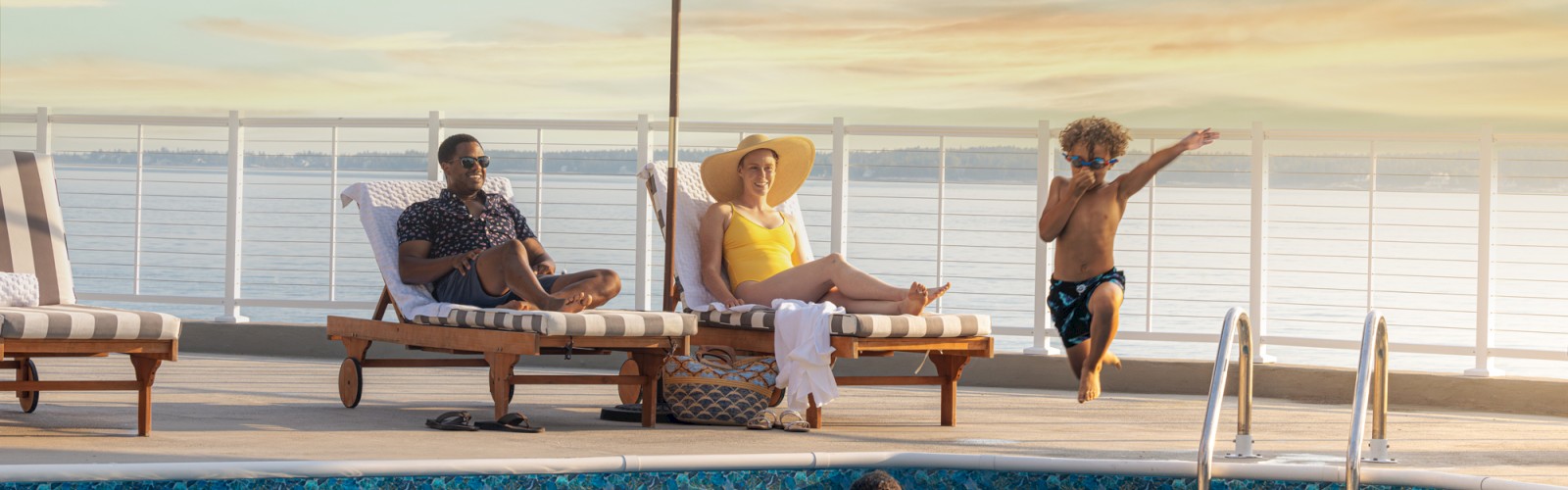 A family relaxing by a pool, with two adults on lounge chairs under umbrellas and two children playing in the water at sunset, is shown.